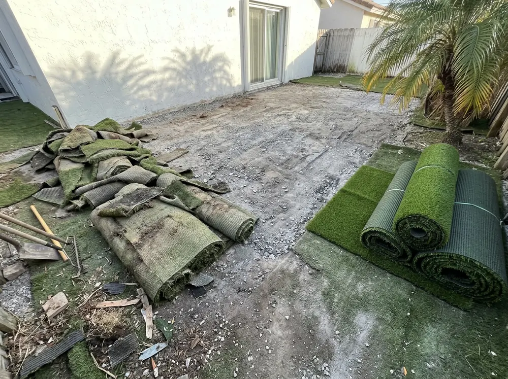 Backyard with faded old artificial turf cut into rolled strips on one side and new turf rolls waiting on the other, the gray stone base exposed between